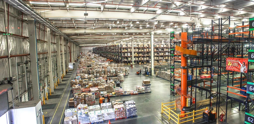A warehouse worker holding a clipboard beside racking, comparing a printed count sheet against boxed stock